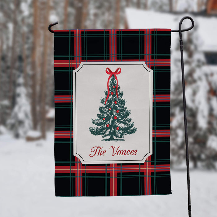 A personalized holiday garden flag hanging from a black metal stand in a snowy outdoor setting. The flag features a black and red plaid pattern with a central white label containing a green Christmas tree illustration and the text 'The Vances'.