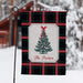 A decorative garden flag hanging from a black metal stand in a snowy outdoor setting. The flag has a black tartan plaid border with red and green stripes, a central cream panel, a green Christmas tree illustration with a red bow, and the text 'The Porters' in red script.
