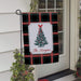A personalized Christmas garden flag hanging from a black metal stand on a front porch. The flag features a black and green plaid background with a central white label containing an illustration of a decorated Christmas tree and the text 'The Morgans'.