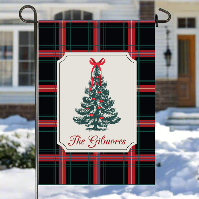A decorative garden flag hanging from a black metal stake outdoors in a snowy setting. The flag has a black plaid pattern with red and green lines, a central cream panel, a green Christmas tree with red ornaments and bow, and the text 'The Gilmores' in red script.