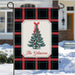 A decorative garden flag hanging from a black metal stake outdoors in a snowy setting. The flag has a black plaid pattern with red and green lines, a central cream panel, a green Christmas tree with red ornaments and bow, and the text 'The Gilmores' in red script.