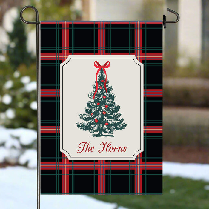 A decorative garden flag hanging from a black metal stake outdoors. The flag has a black, red, and green plaid pattern with a central cream panel showing a green Christmas tree with red ornaments and the text 'The Horns'.