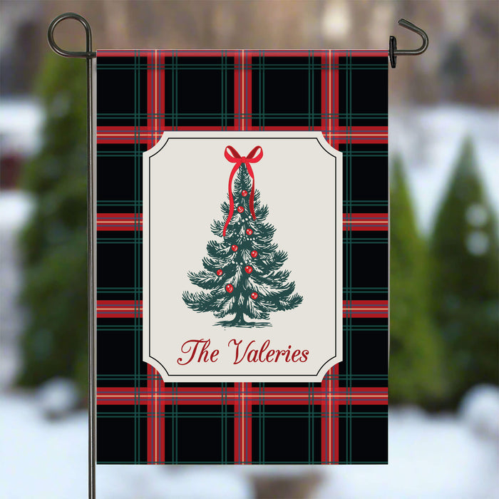 A vertical garden flag with a black, red, and green plaid pattern, featuring a central cream-colored label with an illustration of a decorated Christmas tree and the text 'The Valeries'. The flag is displayed on a black metal stand against a blurred outdoor winter background.