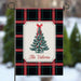 A vertical garden flag with a black, red, and green plaid pattern, featuring a central cream-colored label with an illustration of a decorated Christmas tree and the text 'The Valeries'. The flag is displayed on a black metal stand against a blurred outdoor winter background.