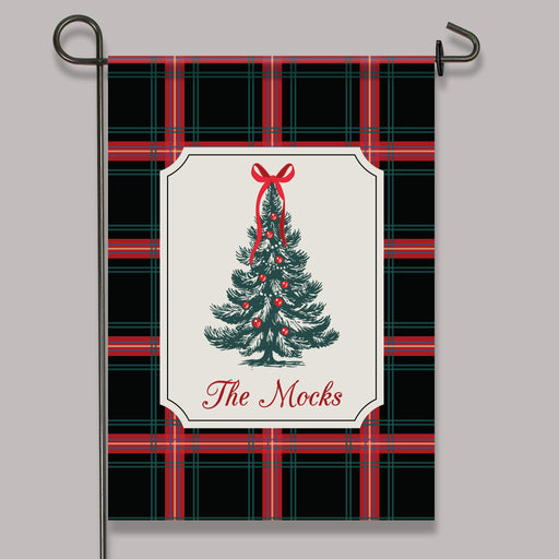 A decorative garden flag hanging from a black metal stand against a light gray background. The flag has a black, red, and green plaid pattern with a central cream label featuring a green Christmas tree illustration, red ornaments, and the text 'The Mocks'.