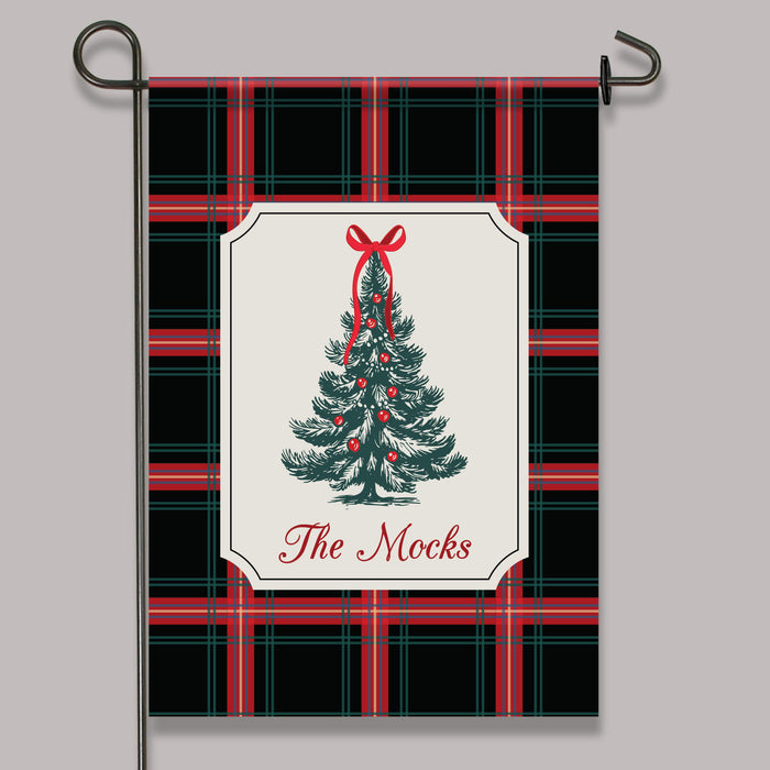 A decorative garden flag hanging from a black metal stand against a light gray background. The flag has a black, red, and green plaid pattern with a central cream label featuring a green Christmas tree illustration, red ornaments, and the text 'The Mocks'.