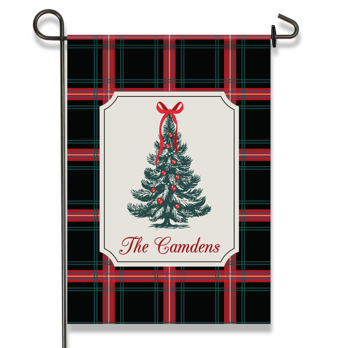 A vertical garden flag on a black metal stand, featuring a black, red, and green plaid pattern with a central cream label showing a decorated Christmas tree and the text 'The Camdens'.