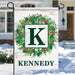 Personalized outdoor garden flag featuring a white wood-textured background with a green wreath, red berries, a large green letter K, and the name KENNEDY at the bottom, displayed on a metal stand in front of a snowy house.