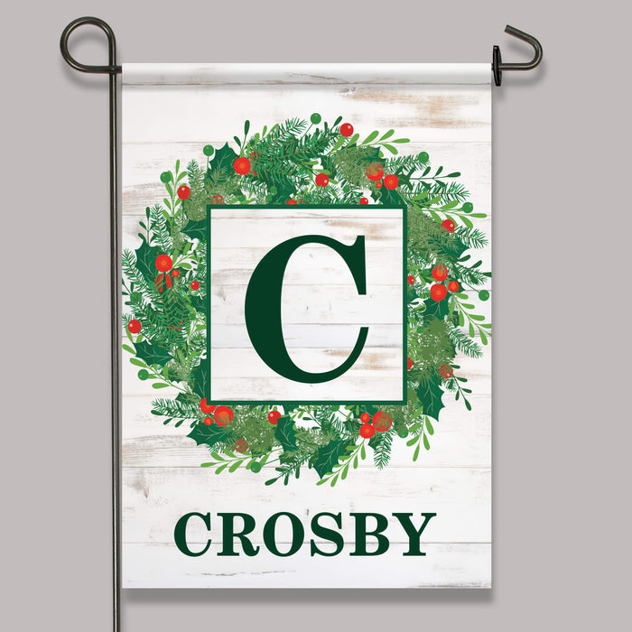 A vertical garden flag hanging from a black metal stand against a light gray background. The flag has a white wood-plank style design with a green wreath, red berries, a large dark green letter C in a white square, and the name CROSBY at the bottom.
