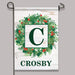 A vertical garden flag hanging from a black metal stand against a light gray background. The flag has a white wood-plank style design with a green wreath, red berries, a large dark green letter C in a white square, and the name CROSBY at the bottom.
