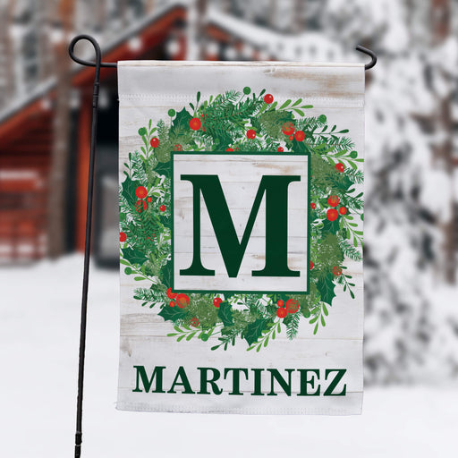 A personalized garden flag with a green wreath, red berries, and a large green letter 'M' on a white wood-textured background, with the name 'MARTINEZ' printed below. The flag is displayed on a black metal stand in a snowy outdoor setting with a blurred cabin in the background.