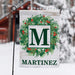 A personalized garden flag with a green wreath, red berries, and a large green letter 'M' on a white wood-textured background, with the name 'MARTINEZ' printed below. The flag is displayed on a black metal stand in a snowy outdoor setting with a blurred cabin in the background.
