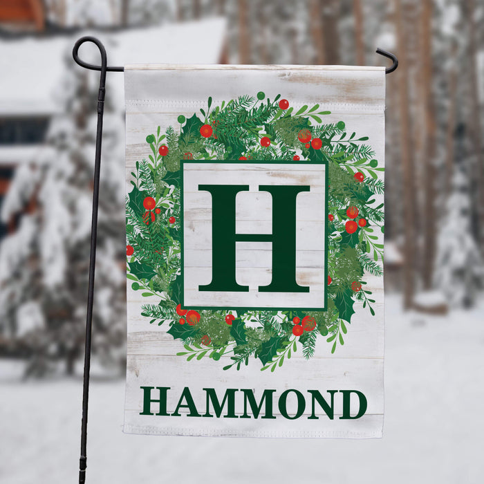A personalized garden flag hanging from a black metal stand in a snowy outdoor setting. The flag features a white wood-textured background with a green wreath decorated with red berries, a large green letter 'H' in a white square, and the name 'HAMMOND' printed at the bottom in green.