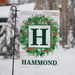 A personalized garden flag hanging from a black metal stand in a snowy outdoor setting. The flag features a white wood-textured background with a green wreath decorated with red berries, a large green letter 'H' in a white square, and the name 'HAMMOND' printed at the bottom in green.