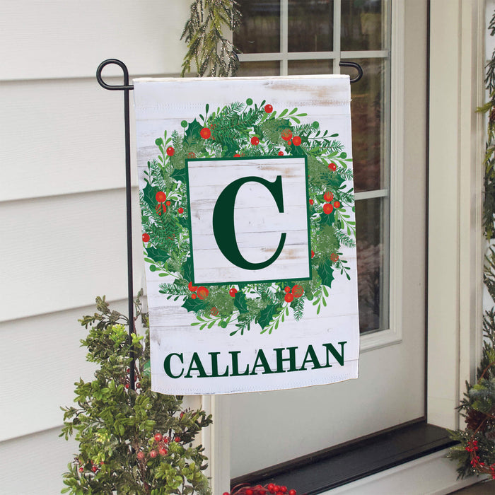 A personalized Christmas garden flag hanging from a black metal stand on a front porch. The flag has a white wood-textured background with a green wreath design, a large green letter 'C' in a white square, and the name 'CALLAHAN' printed at the bottom.