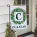 A personalized Christmas garden flag hanging from a black metal stand on a front porch. The flag has a white wood-textured background with a green wreath design, a large green letter 'C' in a white square, and the name 'CALLAHAN' printed at the bottom.