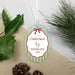 Personalized oval Christmas ornament with a white hanging ribbon, featuring a white label with red border, green and red text reading 'my first CHRISTMAS Hayden Rae 2026,' plus small green baby footprints. Styled on a beige fabric surface with green leaves, pine needles, and pinecones around it.