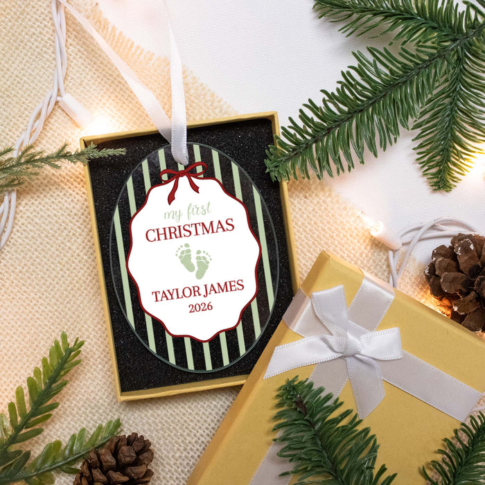 Personalized Christmas ornament in a gift box with green striped edges and a white label that reads 'my first CHRISTMAS TAYLOR JAMES 2026' with baby footprints. Styled flat-lay with pine branches, a gold gift box, and warm string lights.