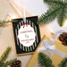 Personalized Christmas ornament in a gift box with green striped edges and a white label that reads 'my first CHRISTMAS TAYLOR JAMES 2026' with baby footprints. Styled flat-lay with pine branches, a gold gift box, and warm string lights.