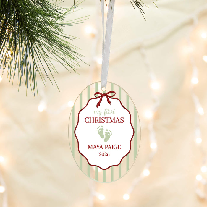 Personalized oval Christmas ornament hanging from a white ribbon, featuring green stripes, red border details, and a custom print with baby footprints and the text 'my first CHRISTMAS MAYA PAIGE 2026'. Pine branches and warm string lights are softly blurred in the background.