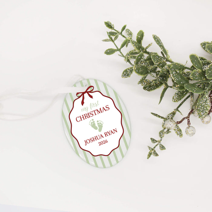 A personalized Christmas ornament with green stripes, featuring a red border, white center, baby footprints, and the text 'my first CHRISTMAS JOSHUA RYAN 2026'. The tag is decorated with a white ribbon and styled next to a green frosted holly branch on a clean white background.