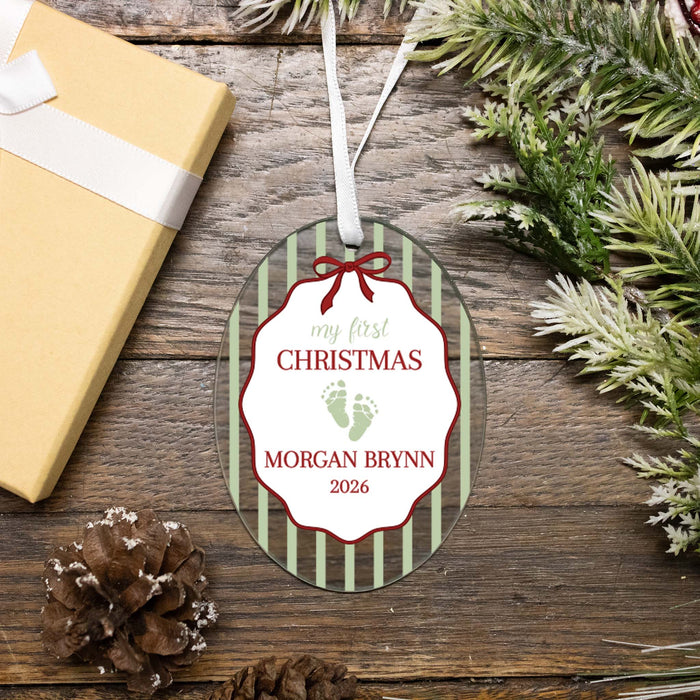 Personalized oval Christmas ornament with a white hanging ribbon, featuring green vertical stripes, a red bow outline, and the text 'my first CHRISTMAS' and 'MORGAN BRYNN 2026' with small green baby footprints. The ornament is photographed on a rustic wooden surface with a wrapped gift box, pinecones, and evergreen branches around it.