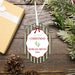 Personalized oval Christmas ornament with a white hanging ribbon, featuring green vertical stripes, a red bow outline, and the text 'my first CHRISTMAS' and 'MORGAN BRYNN 2026' with small green baby footprints. The ornament is photographed on a rustic wooden surface with a wrapped gift box, pinecones, and evergreen branches around it.