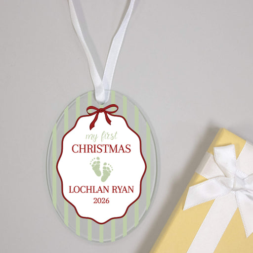 Personalized oval glass Christmas ornament with a white ribbon hanger, featuring a white central label with red border, green and red text reading 'my first CHRISTMAS LOCHLAN RYAN 2026', and green baby footprints. A yellow gift box with a white ribbon is partially visible at the bottom right on a light gray background.