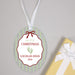 Personalized oval glass Christmas ornament with a white ribbon hanger, featuring a white central label with red border, green and red text reading 'my first CHRISTMAS LOCHLAN RYAN 2026', and green baby footprints. A yellow gift box with a white ribbon is partially visible at the bottom right on a light gray background.