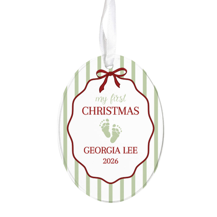 Personalized oval Christmas ornament with a white background, light green vertical stripes, a red scalloped border, a red bow illustration, and a white hanging ribbon. The ornament reads 'my first CHRISTMAS', has baby footprints, and 'GEORGIA LEE 2026'.