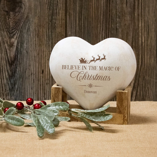 A heart-shaped white 3D heart engraved with 'BELIEVE IN THE MAGIC OF Christmas' and the name 'Donovan', displayed on a small wooden stand with holiday greenery and red berries in front, against a rustic dark wood background.
