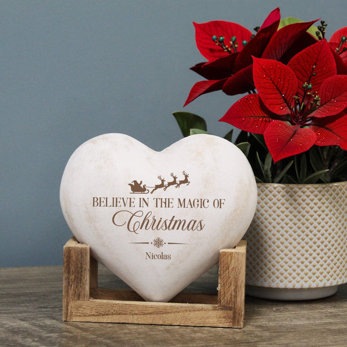 A heart-shaped white Christmas decoration with engraved brown text reading 'Believe in the Magic of Christmas' and the name 'Nicolas', displayed on a small wooden stand. A potted red poinsettia sits behind it against a soft blue-gray wall.