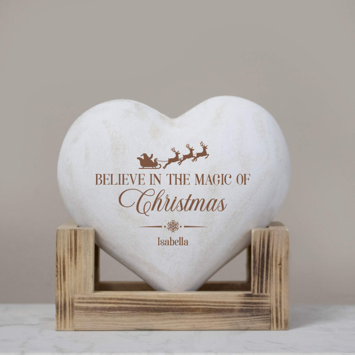 A heart-shaped white wood plaque with a brown engraved Christmas design and the text 'BELIEVE IN THE MAGIC OF Christmas' and the name 'Isabella', displayed on a small wooden stand against a neutral beige background.