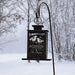 A black metal winter bird feeder hanging from a curved black pole outdoors in a snowy landscape. The front panel has engraved white text reading 'Winter Wishes' and a bird illustration with the phrase 'drift with every song, where little birds have flown all along' and 'The Fleegers.
