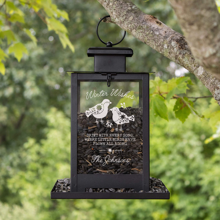 A black metal hanging bird feeder with a clear front panel, filled with sunflower seeds, suspended from a tree branch outdoors. The panel is engraved with bird illustrations and the words 'Winter Wishes' plus a personalized name, with a blurred green forest background.