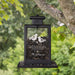 A black metal hanging bird feeder with a clear front panel, filled with sunflower seeds, suspended from a tree branch outdoors. The panel is engraved with bird illustrations and the words 'Winter Wishes' plus a personalized name, with a blurred green forest background.