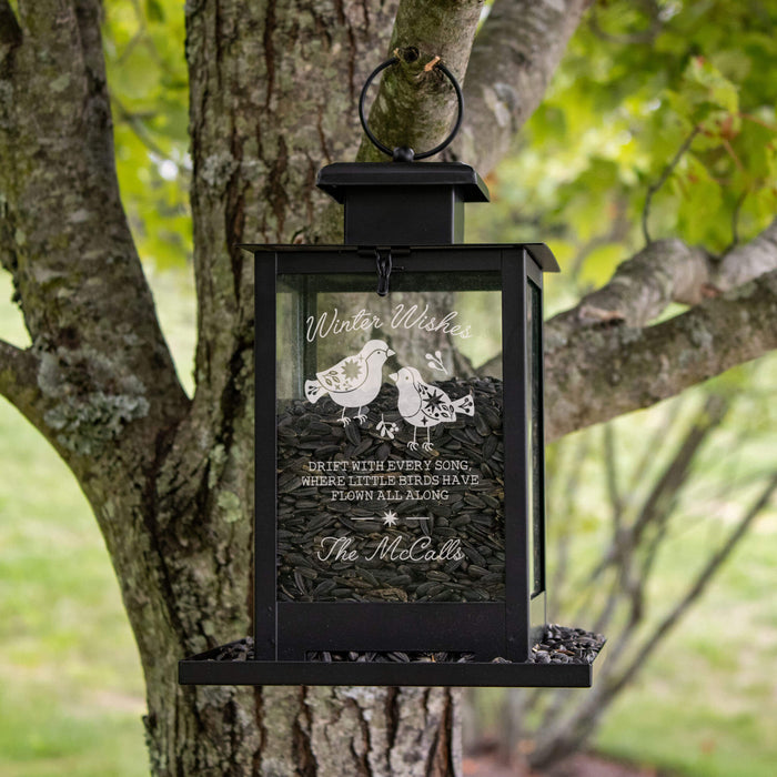 A black metal hanging bird feeder mounted on a tree outdoors. The clear front panel shows two birds and the text 'Winter Wishes' and a winter poem, with dark seed fill visible inside.