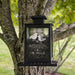 A black metal hanging bird feeder mounted on a tree outdoors. The clear front panel shows two birds and the text 'Winter Wishes' and a winter poem, with dark seed fill visible inside.