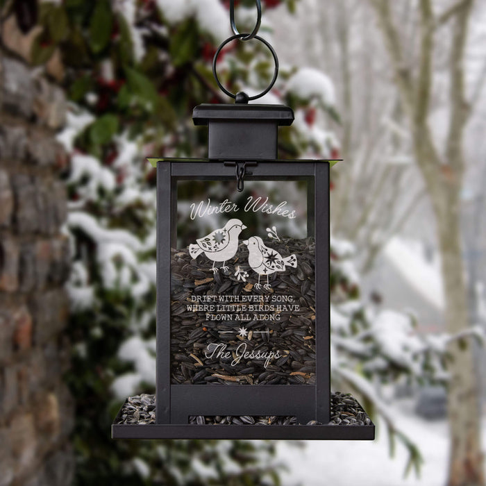 A black metal hanging bird feeder lantern filled with sunflower kernels, with a clear front panel engraved with the words 'Winter Wishes' and an illustration of two birds, set against a blurred snowy garden background.