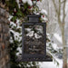 A black metal hanging bird feeder lantern filled with sunflower kernels, with a clear front panel engraved with the words 'Winter Wishes' and an illustration of two birds, set against a blurred snowy garden background.