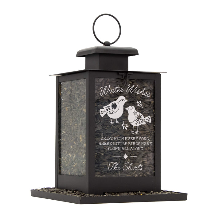 A black metal bird feeder with a hanging loop and a base filled with seed. The front panel has engraved text reading 'Winter Wishes' and 'The Shorts,' plus two bird illustrations.