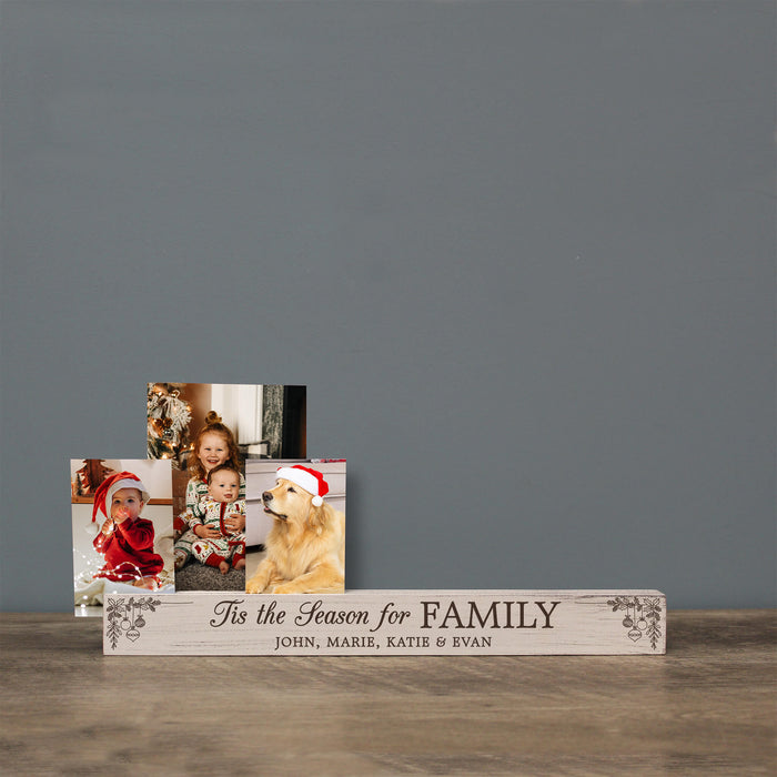 A personalized wooden family photo stand on a rustic wooden table against a solid gray wall. The stand is rustic wood with dark brown engraved text reading 'Tis the Season for FAMILY' and 'JOHN, MARIE, KATIE & EVAN'.