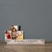A personalized wooden family photo stand on a rustic wooden table against a solid gray wall. The stand is rustic wood with dark brown engraved text reading 'Tis the Season for FAMILY' and 'JOHN, MARIE, KATIE & EVAN'.