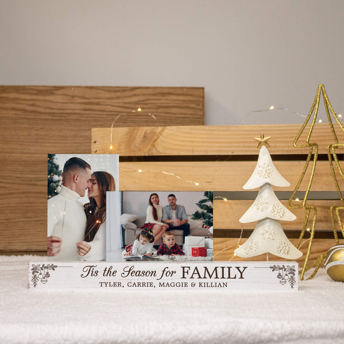 A personalized wooden Christmas photo frame stand with two printed family photos and the text 'Tis the Season for FAMILY' and names below, styled on a white surface with a wooden crate, string lights, a white ceramic Christmas tree, and gold wire decor in the background.