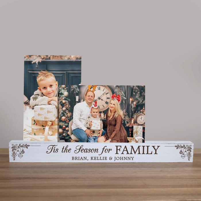A personalized wooden Christmas photo display bar on a tabletop, featuring two printed holiday family photos stacked above custom text that reads 'Tis the Season for FAMILY' and 'BRIAN, KELLIE & JOHNNY'. The sign itself is white with dark brown engraved lettering and small decorative ornament motifs at the corners.