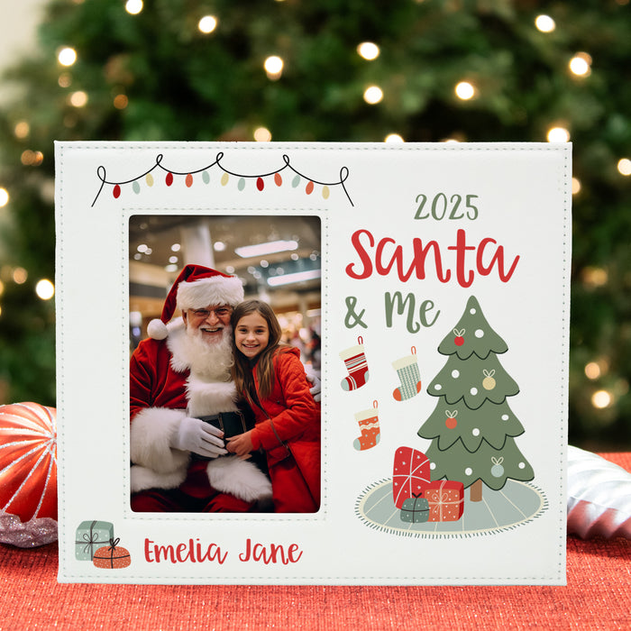 A personalized white Christmas photo frame displayed on a red surface with a blurred Christmas tree in the background. The frame features a photo of Santa and a young girl, along with the text '2025 Santa & Me' and the name 'Emelia Jane', decorated with illustrations of a Christmas tree, stockings, and gifts.