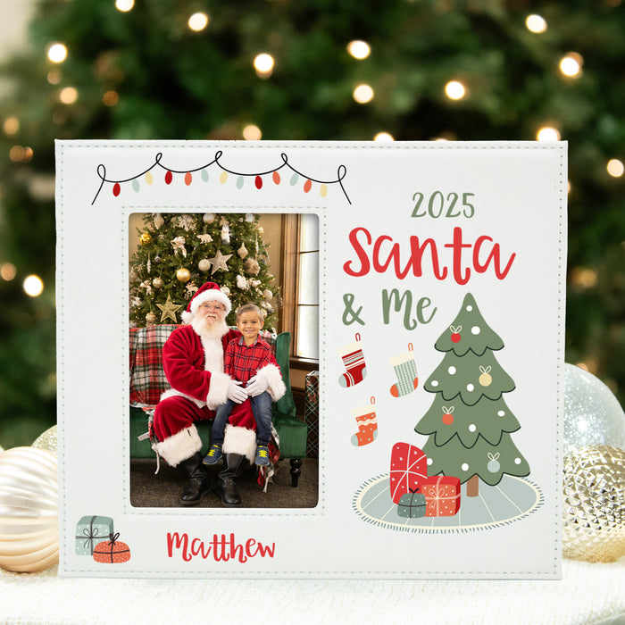 A personalized white Christmas photo frame with a stitched border, featuring a custom photo of Santa and a child, the text '2025 Santa & Me' and 'Matthew', a green Christmas tree illustration, and holiday decorations. The frame is displayed on a white surface with a blurred festive background of a Christmas tree and ornaments.