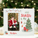 A personalized white Christmas photo frame with a stitched border, featuring a custom photo of Santa and a child, the text '2025 Santa & Me' and 'Matthew', a green Christmas tree illustration, and holiday decorations. The frame is displayed on a white surface with a blurred festive background of a Christmas tree and ornaments.