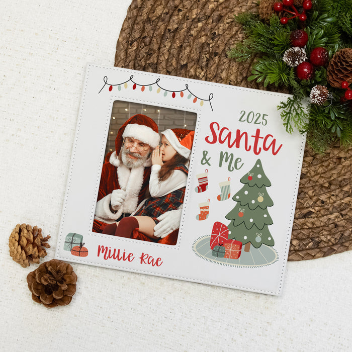 A white personalized Christmas photo frame with decorative holiday graphics, shown on a white textured surface with pinecones, a woven mat, and greenery. The frame includes a photo slot with a Santa and child image, the text '2025 Santa & Me', and the name 'Miliie Rae' in red script.