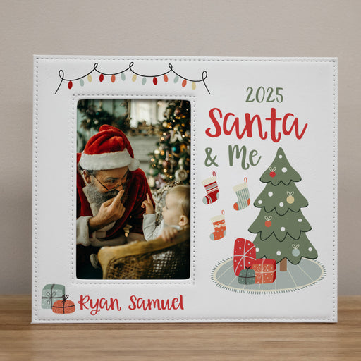A personalized white Christmas photo frame with a stitched border, featuring a customizable photo slot, the text '2025 Santa & Me', a green Christmas tree with ornaments and presents, and the name 'Ryan Samuel' in red script. The frame is shown standing on a wooden surface against a neutral beige wall.